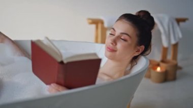 Banyo yapan kadın, modern ev tuvaletinde kitap okuyor. Köpük banyosunda yatan hayalperest kadın beyaz köpükle kaplanmış romantik romanların keyfini çıkarıyor. Lüks küvette elinde edebiyatla sırılsıklam sırılsıklam gülümseyen kız..