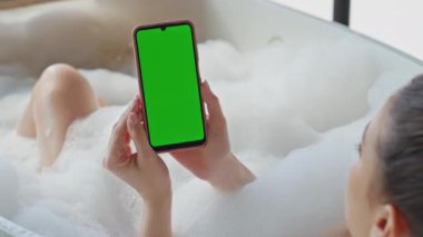 Küvette içi köpüklerle dolu yeşil ekran akıllı telefonlu banyo yapan bir kadın. Krom telefon tutan kadın sakin anların tadını çıkarıyor. Huzurlu kadın banyoda cep telefonuyla sosyal medyada geziniyor..