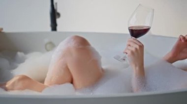 Esmer, lüks banyoda şarap banyosuyla banyo yapıyor. Güler yüzlü kadın elinde cam bardak, elinde beyaz köpükle akıllı telefondan konuşuyor. Mutlu bayan vücut bakımını sever..