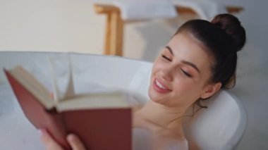 Köpük banyosunda okuyan kız, kişisel bakım rutinini rahatlatmanın keyfini çıkarıyor. Gülümseyen kadın spa küvetinde beyaz köpükle sırılsıklam kitap tutuyor. Lüks banyoda yatan edebiyatla ilgilenen mutlu esmer..