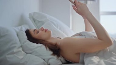Yatak odasında akıllı telefon kullanan esmer, sabah yakın planda uyanmış. Sakin bir kadın beyaz yatak yastığında rahat rahat cep telefonuyla sosyal medyada geziniyor. Mutlu kız yatakta uzanıyor Tembel hafta sonunun tadını telefonla çıkarıyor.