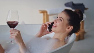 Küvette gevşeyen ve şarap kadehini yakın plan tutan telekız. Neşeli kadın, spa köpüğü banyosunda elinde alkol kadehiyle uzanıp akıllı telefonuyla konuşuyor. Gülümseyen esmer, köpüklü, konuşan cep telefonlu..