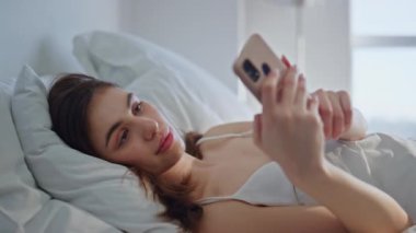Yatakta mesaj yazan, rahat beyaz yataklarda rahatlayan bir kız. Sakin esmer mesajcı akıllı telefon yastık kılıfı yatak odasında yatıyor. Huzurlu kadın çevrimiçi iletişimden zevk alıyor..