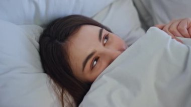Yataktan gizlice bakan neşeli bir kadın neşe dolu bir yakınlaşma hissediyor. Beyaz battaniyenin altında saklanan tatlı, güzel esmer bir kız. Sıcak bir evde uyanıyor. Mutlu genç bayan rahat yatak odasında pozitif sabahın tadını çıkarıyor..
