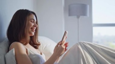Yatakta cep telefonuyla mesaj atan gülümseyen kadın. Neşeli genç bir kadın, rahat bir yatakta uyanan akıllı telefon mesajı okuyor. Mutlu rahat kız, sabahın erken saatlerinde evde cep telefonuna bakıyor..