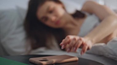 Uykulu kız güneşli bir sabah yatakta akıllı telefona uzanıyor. Esmer, rahat bir yatak odasında uyanıyor cep telefonunun alarmını kapatıyor ve yorgun hissediyor. Uykusuz bir kadın. Uykulu bir ifadeyle telefona bakıyor..