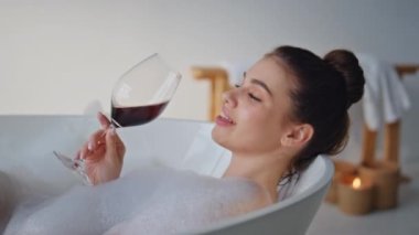 Köpük banyosunda şaraptan zevk alan gülümseyen kadın. Şarap kadehinden alkol içen dingin kadın küvette beyaz köpükle dinleniyor. Banyoda rahat bir şekilde gülümseyen mutlu esmer..