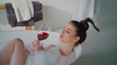 Şarap banyosu yapan rahat bir kadın. Huzur verici bir yakınlaşma. Banyoda bir bardak Merlot ile köpük banyosunun keyfini çıkaran mutlu bayan. Beyaz köpükle kaplıca küvetinde kırmızı alkol içen sakin bir kız..