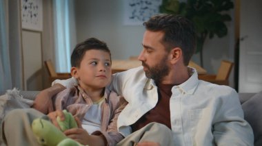 Baba oğlunun akşam oturma odasından konuşmasını kucaklıyor. Bekar ebeveyn, küçük çocuk rahat koltukta birlikte dinleniyor. Sevimli bebek elinde yumuşak oyuncakla babacığıyla eğleniyor. Çocukluk kavramı