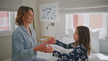 Işıklı yatak odasında birlikte oynayan pozitif aile. Neşeli anne küçük çocuk el çırpıyor beyaz yatakta eğleniyor. Gülen kadın küçük kız, düşen yastıklar, keyifli bir daire.