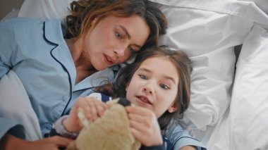 Anne yeni yürümeye başlayan çocuk, güzel aydınlatılmış yatak odasında yatarken eğleniyor. Pijamalı rahat bayan küçük çocuk yatakta oyuncak ayıyla oynamaktan zevk alıyor. Sakin anne kızı beyaz çarşaflarda dinleniyor. 