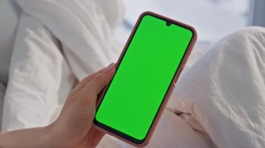 Açık ev yatak odasındaki yeşil ekranlı cep telefonuna dokunuyor. Kimliği belirsiz bir kadın, beyaz bir yatakta krom anahtarlı akıllı telefon tutuyor. Dairede gezen kız model cep telefonu uygulaması.