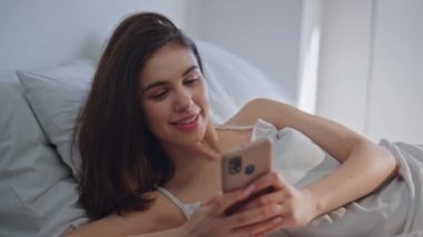 Sabahın erken saatlerinde rahat bir yatakta akıllı telefondan mesaj okuyan mutlu bir kadın. Neşeli esmer, rahat yastıklarda uzanan cep telefonu görünümlü. Yatak odasında uyanan mutlu bayanın cep telefonu mesajları..