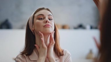 Banyo aynasında cilt bakımı yapan güzel bir kadın. Yüzünde sakin bir ifadeyle boynuna masaj yapan mutlu bayan yansımaya bakıyor. Evde doğal güzelliği olan zarif bir kız portresi..