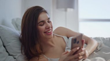 Sabahın erken saatlerinde yatağın üzerine akıllı telefon mesajı atan bir kız. Beyaz yatak yastıklarında uzanan neşeli kadın cep telefonu ekranına bakıyor. Neşeli esmer, güneşli yatak odasında çevrimiçi etkileşimden hoşlanıyor..