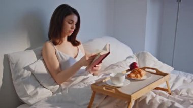 Sakin esmer kadın kitap okuyor, yatakta kahvaltı yapıyor. Huzurlu bir kadın, en sevdiği romanla sakin bir sabah geçiriyor. Yatak odasında edebiyat hobisiyle rahatlayan rahat bir bayan..