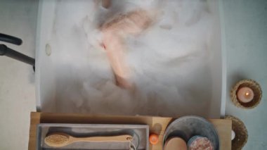 Güzel bir kadın küvete girmiş. Beyaz tüylü köpüklerle dolu. Huzurlu bir üst manzara. Sakin bir bayan mumlarla spa gününü rahatlatıyor. Meditasyon yapan kız sabun köpükleriyle sağlıklı ciltleri nazikçe yıkıyor.
