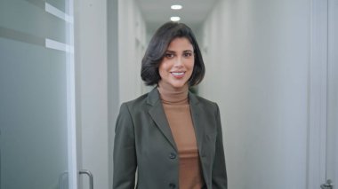 Şirket koridorunda poz veren kendine güvenen bayan patron. Modern ofis ortamında gülümseyen zarif iş kadınının portresi. Profesyonel çevrede pozitif enerji saçan mutlu bir kadın.