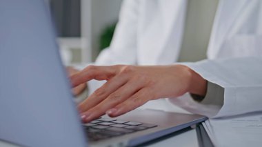 Terapist elleri laptopta tıbbi veri girişi yapıyor. Modern klinik ofisinde oturuyorlar. Hastanede daktilo kullanan odaklanmış kadın doktor. Beyaz cüppeli doktor idari iş raporlarıyla meşgul..