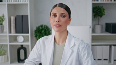 Modern klinik ortamında poz veren kendine güvenen bir doktor. Hastanenin içinde beyaz önlüklü kadın doktor portresi. Poliklinikteki kamerada gülümseyen dost canlısı sağlık uzmanı.