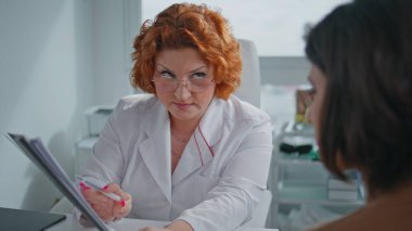 Sağlık danışmanı klinikteki kadın hastaya sigorta poliçesi gösteriyor. Kızıl saçlı doktor konsültasyonda tıbbi prosedür öneriyor. Sağlık sigortası belgelerini açıklayan gözlüklü bir profesyonel.