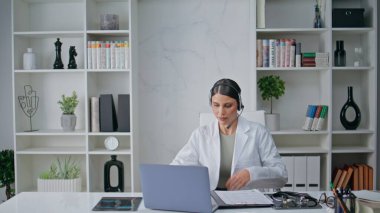 Doctor start online meeting wearing headset in modern clinic interior closeup. Positive woman medic sitting at laptop join to medical educational virtual event. Smiling physician work at telemedicine clipart