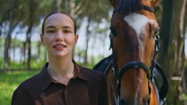 Güneşli parkta atıyla yürüyen neşeli kovboy kız portresi. Gülümseyen kadın, elinde evcil hayvan tasmasıyla doğada birlikte eğleniyor. Neşeli kadın, tapılası bir hayvanla poz veriyor yeşil ortamda.