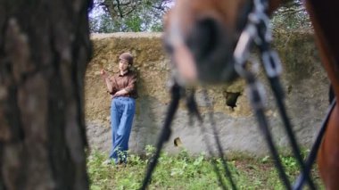 Çiftlik bölgesinde duvara yaslanan romantik bir atlı kadın. Vahşi kahverengi at ilk planda ayakta duruyor. Atlı kadın elinde çivili kamera ile gün ışığına bakıyor. Kendinden emin jokey taşlanmış çit pozu veriyor.