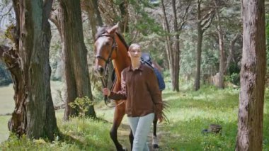 Young woman strolling with horse in green park. Relaxed girl looking at adorable brown animal enjoying peaceful nature. Calm pet stepping on grass with affectionate female owner at summer outdoors