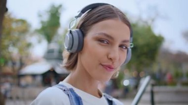 Güneşli bir sokakta kulaklıkla poz veren bir kadın. Neşeli kız kulaklıkla müzik dinliyor kameraya bakıyor ve gülümsüyor. Olumlu gülümseyen hipster modern dijital cihazda sesin tadını çıkarıyor.