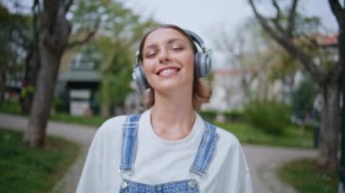Gezgin bir kadın, kablosuz kulaklıkla müzik dinliyor. Güzel yeşil parkın yakın çekiminde. Modern kulaklıklarla gezen neşeli görünümlü bir kız. Gülümseyen bayan hippi haftasonu tatilinin tadını çıkar.