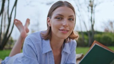 Parktaki çimlerin üzerinde uzanmış romantizm okuyan gülümseyen kız. Tasasız, güzel bir bayan, yeşil çimenlikte elinde kitapla kameraya bakıyor. Mutlu kadın dinleniyor ve doğa üzerinde çalışıyor. Pozitif esmer, hobi sahibi..