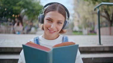Hafta sonları Sunny Park 'ta oturup kitap okuyan gülen kadın. Neşeli kız şehir caddesinde eğlence edebiyatının tadını çıkarıyor. Mutlu gülümseyen kadın kulaklıkla müzik dinliyor kameraya bakıyor