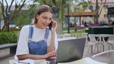 Akıllı telefondan konuşan kız açık kafede dizüstü bilgisayar görüntüsü alıyor. Gülümseyen kadın cep telefonunu arıyor Green Park Terasında bilgisayarla çalışıyor. Akıllı kadın telefonda pazarlık yapıyor.