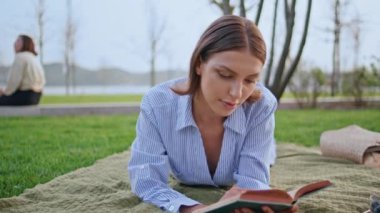 Huzurlu parkta uzanan çimleri okuyan bir kadın. Yakın plan. Mavi elbiseli romantik bayan piknik battaniyesinde ilginç bir kitabın keyfini çıkarıyor. Rahat öğrenci doğa sükunetinde edebiyata odaklanıyor.