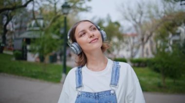 Kulaklıklı genç caddede yürüyen güzel yaz gününün tadını çıkarıyor. Green Park 'ta kablosuz kulaklıkla müzik dinleyen mutlu kadın. Rahatlamış kız, yeşil doğada geziniyor, neşeli bir gülümsemeyle.