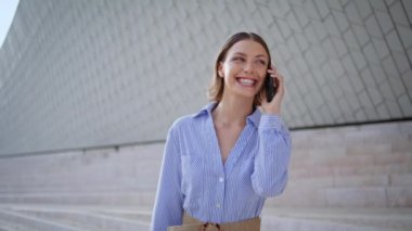 Modern kasaba mimarisinin yakınında cep telefonuyla konuşan mutlu bir kadın. Gülümseyen bayan şehir caddesinde akıllı telefon konuşmasından hoşlanıyor. Neşeli kadın esmer, dışarıdaki cep telefonundan neşe saçıyor.