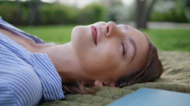 Huzurlu bir ifadeyle piknik battaniyesine uzanmış bir kadın. Kaygısız güzel kız huzur parkındaki yeşil çimlerde dinleniyor. Mutlu kadın esmer rahatlamanın keyfini çıkarıyor. Mavi gökyüzüne bakıyor..