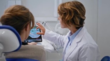 Kadın ortodontist, röntgeni stomatoloji kliniğinde tablet kullanarak açıklıyor. Gülümseyen dişçi danışmada dijital oral bakım gösteriyor. Stomatolog hastayla tedavi seçeneklerini tartışıyor