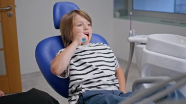 Stomatoloji kliniğinde dişlerini fırçalayan çocuk diş hekimi. Gülümseyen çocuk modern diş hekimliğinde ağız temizliğine özen gösteriyor. Sevimli küçük hasta dişçi randevusunda sağlık prosedürünün tadını çıkarıyor..