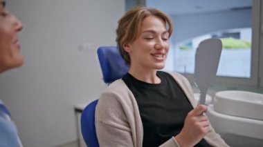 Konuşan neşeli kız dişçi modern klinikte ortodontik prosedürü tamamladı. Mutlu kadın aynaya bakıyor diş tedavisinden sonra gülümsüyor. Profesyonel stomatolog danışmanlığı.