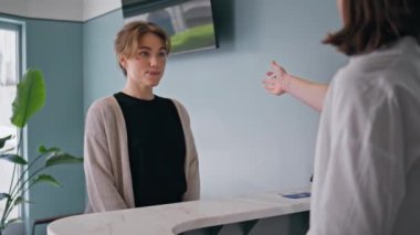 Kadın hasta, sağlık hizmetleri randevusunda resepsiyonistle konuşuyor. Gülümseyen klinik müşterisi resepsiyondaki tıbbi personelle etkileşim halinde. Arkadaş canlısı iletişim ziyaretçisi ve sağlık hizmeti sağlayıcısı.