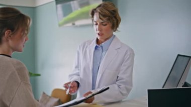 Modern klinikte gülümseyen kadın hastalara belgeleri imzalatmak için çalışan bir sağlık görevlisi. Kadın doktor panoda tedavi planını açıklıyor. Genç ziyaretçi randevu formunu imzalıyor..