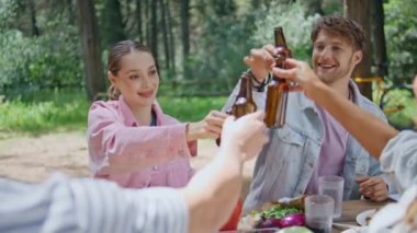 Grup arkadaşları ormanda piknikte şişe kızartıyorlar. Neşeli yaz toplantısında mutlu insanlar birlikte kutluyorlar. Gülümseyen gençler bira içip hafta sonunun tadını çıkarıyorlar. Doğayla iç içe.