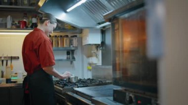 Genç aşçı kaşıkla tepsiyi karıştırıp ocakta yemek hazırlıyor. Profesyonel mutfak malzemesi pişirme sosuyla yemek pişiren gülümseyen kadın. Mutlu kafe çalışanı modern mutfağın aşçılık sürecinin tadını çıkarıyor.