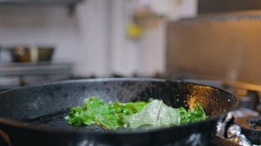 Restoran işçisi modern mutfakta tavada sebze kızartıyor. Bilinmeyen aşçı profesyoneli ısıtmalı fırının üzerinde taze marul hazırlıyor. Aşçı tuzlama malzemeleri mutfak işyerinde pişiyor..