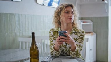 Cafe ziyaretçisi akıllı telefondan mesaj atıyor. İçeride rahat rahat oturuyormuş. Yakın çekim. Kıvırcık saçlı kadın kafeteryada dostça bir toplantı bekliyor. Rahat bir bayan cep telefonundan sosyal medya içeriğini izliyor..