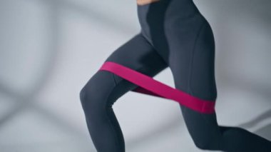 Sportswoman legs squats workout with rubber resistance band at empty space. Unrecognizable woman training butt and hamstring at white wall background. Activating muscles of buttocks thighs concept 