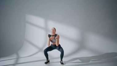 Beyaz boş stüdyoda ağır halterleri kaldıran bir kadın. Sportif bayan spor malzemeleriyle el sıkışıyor. Sağlıklı vücut için spor giyerek egzersiz yapan formda genç bir manken. Aktif spor yaşam tarzı