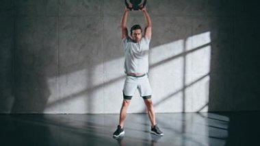 Çatı katında ağırlık çalışması yapan sporcu bir adam. Güçlü güçlü esmer, ağır ekipman kaldırıyor gri duvar arka planında kollarını kaldırıyor. Spor kulübünde tek başına omuz çalışması yapan ciddi bir sporcu. 